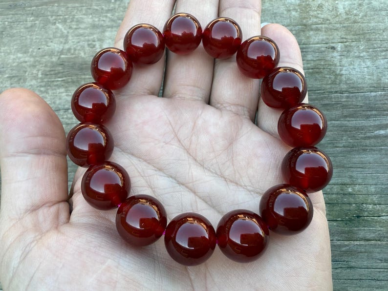 May include: A bracelet featuring large, polished, round beads in a deep, rich red color. The beads are strung together, forming a circular shape. The bracelet is displayed on a hand, showcasing the jewelry's size and color.
