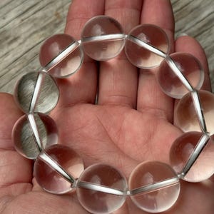 May include: A circular bracelet composed of eleven smooth, clear, spherical beads. Each bead is transparent and polished, reflecting light on its surface. The beads are strung together in a continuous loop, creating a uniform and symmetrical design.
