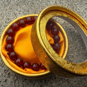 May include: A beaded bracelet with dark red beads rests inside a round, gold-colored box. The box is lined with yellow fabric. The lid of the box is open, revealing the bracelet and the interior.