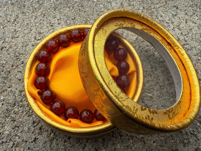 May include: A beaded bracelet with dark red beads rests inside a round, gold-colored box. The box is lined with yellow fabric. The lid of the box is open, revealing the bracelet and the interior.