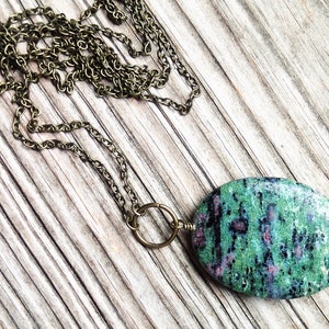 May include: A green and pink oval stone pendant necklace with a brass chain. The stone has a mottled pattern of green, pink, and black.