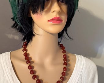 May include: A beaded necklace featuring large, round, dark red beads. The beads are separated by small, gold-colored spacers. The necklace is displayed on a mannequin wearing a white shirt.