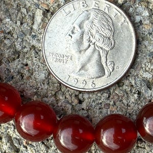May include: A close-up of a quarter coin next to a strand of red-orange gemstone beads. The coin is silver and shows the profile of George Washington. The beads are round and have a glossy finish.