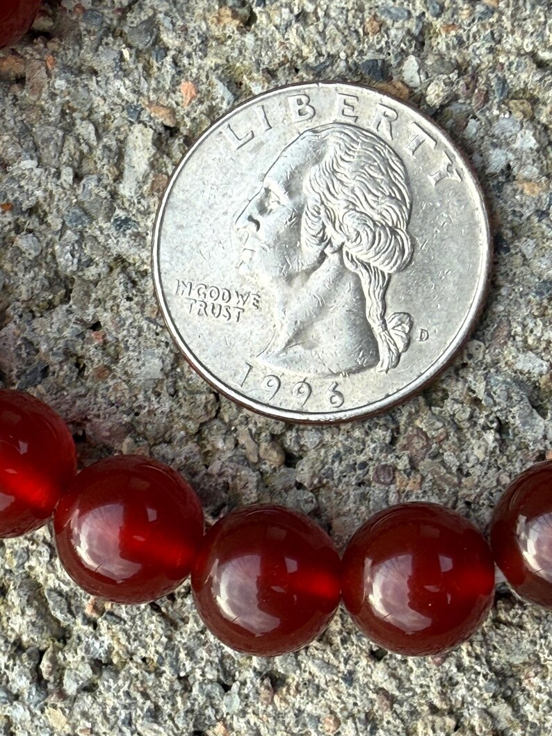 May include: A close-up of a quarter coin next to a strand of red-orange gemstone beads. The coin is silver and shows the profile of George Washington. The beads are round and have a glossy finish.