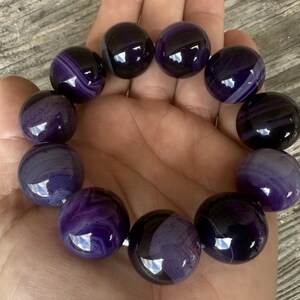 May include: A bracelet made of large, round, polished beads. The beads are a deep purple color with swirling patterns of lighter purple and white. The bracelet is held in a hand, showcasing the smooth, reflective surfaces of the beads.