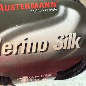 May include: A close-up of a ball of dark gray Merino Silk yarn. The label reads "AUSTERNANN fashion & style" in red. The text "Merino Silk" is prominently displayed in white. The label also indicates a length of approximately 115 meters and a weight of 50 grams.