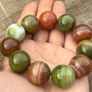 May include: A bracelet of polished, round gemstone beads in shades of green, brown, orange, and white. The beads are held in a hand, showcasing their varied colors and patterns. The bracelet is a natural stone jewelry item.