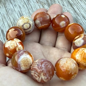 May include: A circular bracelet composed of eleven polished, spherical beads. The beads feature a marbled pattern with varying shades of burnt orange, cream, and deep brown, displaying natural concentric rings and organic, swirling textures.