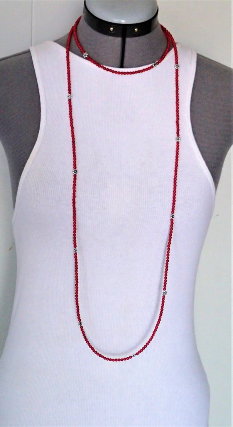 Puede incluir: Un collar largo de cuentas rojas con detalles en forma de flor plateados. El collar est&aacute; superpuesto dos veces, creando una longitud m&aacute;s larga.