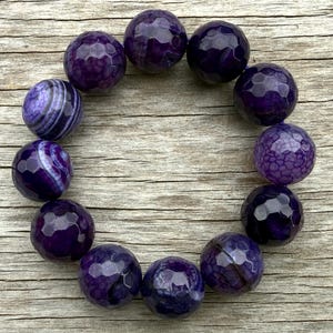 May include: A collection of faceted, deep purple gemstone beads arranged in a circular shape. The beads have a polished, reflective surface, with variations in color and pattern. The background is a light brown wooden surface.