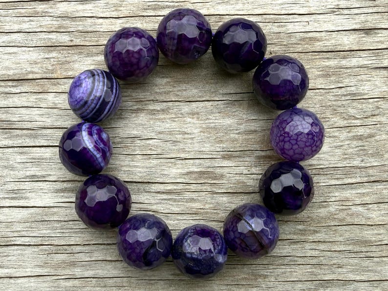May include: A collection of faceted, deep purple gemstone beads arranged in a circular shape. The beads have a polished, reflective surface, with variations in color and pattern. The background is a light brown wooden surface.