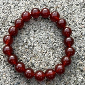 May include: A round bracelet made of polished, deep red beads. The beads are evenly sized and strung together, creating a continuous loop. The bracelet is displayed on a textured, gray surface.