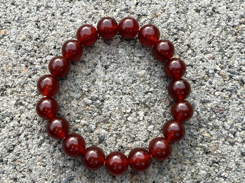 May include: A round bracelet made of polished, deep red beads. The beads are evenly sized and strung together, creating a continuous loop. The bracelet is displayed on a textured, gray surface.