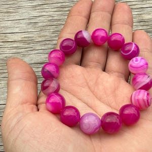 May include: A magenta beaded bracelet with white and pink swirls. The bracelet is made of round, polished beads. The bracelet is held in a hand, with the wooden surface in the background.