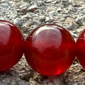 May include: Close-up of three large, glossy, red beads. The beads are perfectly round and reflect light, creating highlights. The beads are resting on a rough, gray stone surface. The beads appear to be part of a necklace or bracelet.