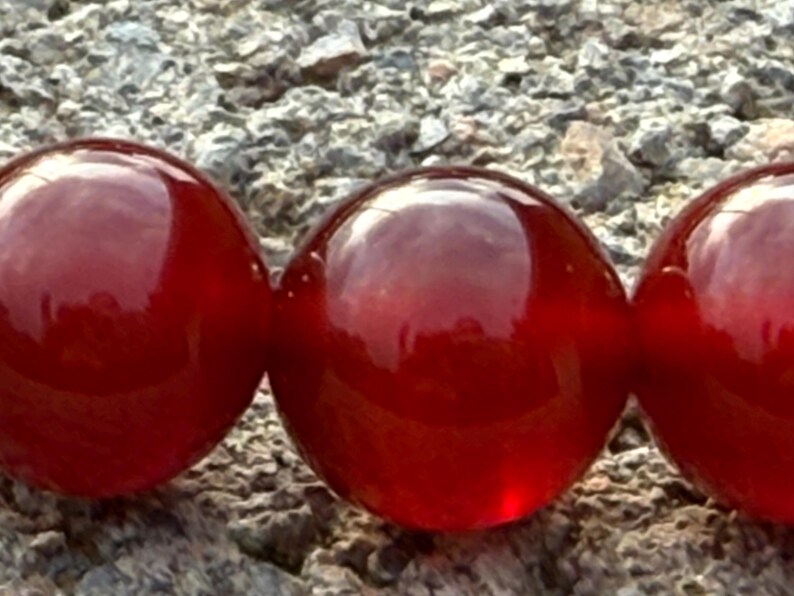May include: Close-up of three large, glossy, red beads. The beads are perfectly round and reflect light, creating highlights. The beads are resting on a rough, gray stone surface. The beads appear to be part of a necklace or bracelet.