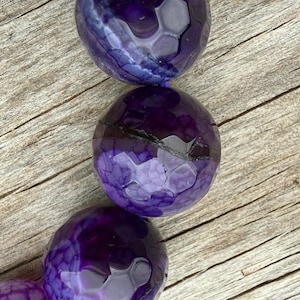 May include: Close-up of a strand of large, round beads in shades of purple. The beads have a textured, faceted surface, and are arranged against a weathered, light brown wooden background. The beads are a deep, rich purple color.