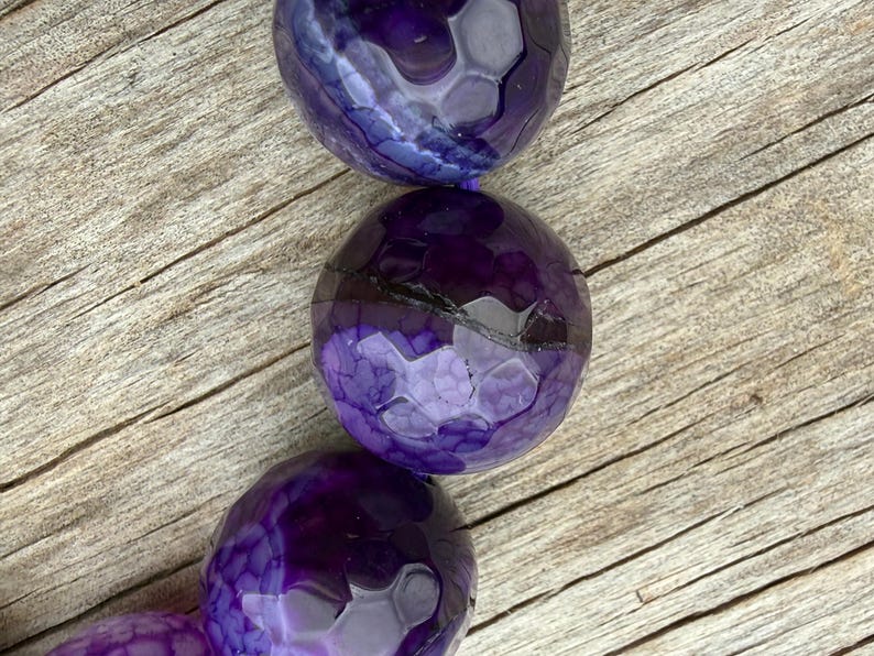 May include: Close-up of a strand of large, round beads in shades of purple. The beads have a textured, faceted surface, and are arranged against a weathered, light brown wooden background. The beads are a deep, rich purple color.