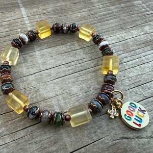 May include: A beaded bracelet featuring yellow, green, and brown beads, with a gold-colored charm that says "GOOD LUCK". The bracelet has a mix of square and round beads.