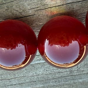 May include: Close-up of a strand of large, round, glossy beads. The beads are a deep, rich red color, reflecting light and the surrounding environment. The beads are strung together, suggesting a necklace or bracelet.
