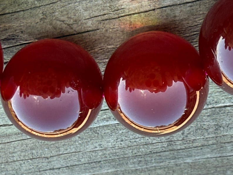 May include: Close-up of a strand of large, round, glossy beads. The beads are a deep, rich red color, reflecting light and the surrounding environment. The beads are strung together, suggesting a necklace or bracelet.