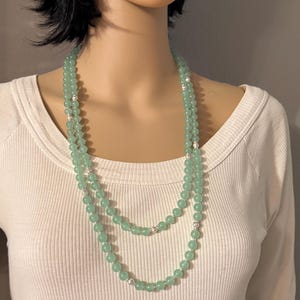 May include: A double-strand necklace featuring light green, round beads. The necklace has silver-colored accents and is displayed on a mannequin wearing a white long-sleeved shirt. The beads appear to be approximately 0.3 inches in diameter.