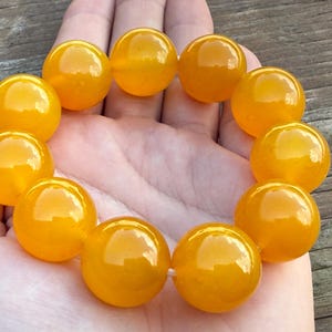 May include: A bracelet of large, round, polished amber-colored beads. The beads are strung together, creating a circular shape. The bracelet is held in a person's hand, with the beads reflecting light and appearing smooth and glossy.