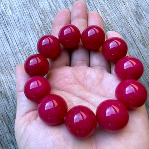 May include: A stretch bracelet composed of twelve large, smooth, spherical beads in a vibrant, glossy magenta-red color. The beads are uniform in size and shape, featuring a polished finish that reflects light, arranged in a circular loop pattern.