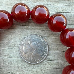 May include: Close-up of a strand of large, glossy, round, dark red beads. A U.S. quarter coin is placed in the center for size comparison. The beads are strung on a pink thread, and the background is a weathered wooden surface.