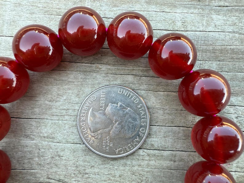 May include: Close-up of a strand of large, glossy, round, dark red beads. A U.S. quarter coin is placed in the center for size comparison. The beads are strung on a pink thread, and the background is a weathered wooden surface.