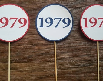 May include: Three round white paper signs with red and blue borders and the year 1979 printed in red on each sign. The signs are attached to wooden sticks.