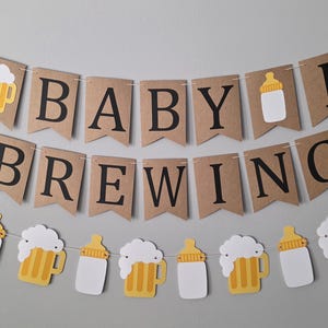 May include: A two-tiered banner set made of brown cardstock pennants. The top tier spells A BABY IS BREWING in black letters, interspersed with a beer mug and baby bottle icon. The bottom tier features alternating beer mug and baby bottle cutouts.