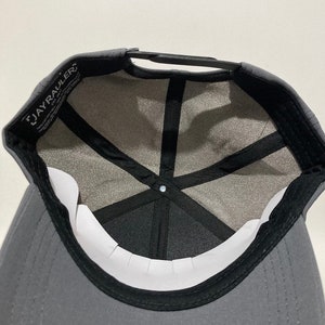 May include: A grey baseball cap with a black brim and a black mesh lining. The cap has a black adjustable strap and a white label with the text "JAYRAULER" on the front.