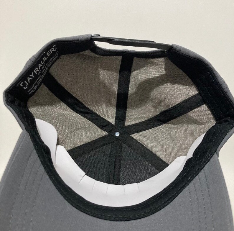 May include: A grey baseball cap with a black brim and a black mesh lining. The cap has a black adjustable strap and a white label with the text "JAYRAULER" on the front.