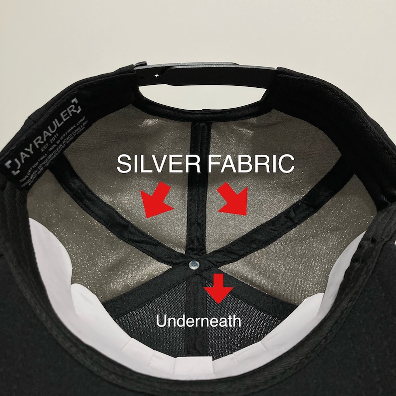 May include: Inside view of a black baseball cap with a white underside. The cap has a black strap and a silver fabric lining. The text "SILVER FABRIC" is printed on the lining, with arrows pointing to the fabric. The text "Underneath" is printed below the arrows.