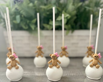 May include: White cake pops topped with small teddy bear decorations wearing pink bows. The cake pops are arranged on a gray surface, with a blurred background of greenery and a white planter.