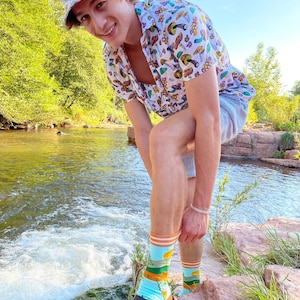 May include: A person wearing a white button-down shirt with a mushroom print, a bucket hat with a mushroom print, and blue and green socks with a mushroom print. The person is standing in a river with rocks and trees in the background.