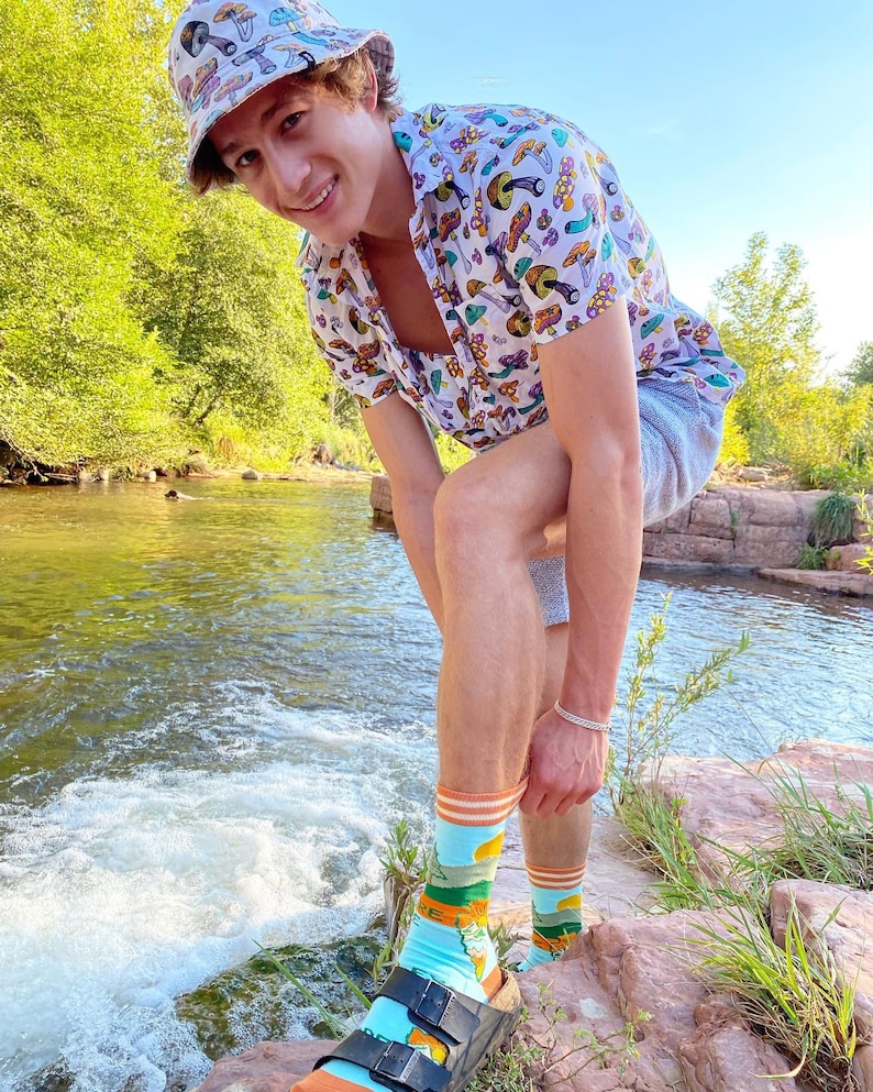 May include: A person wearing a white button-down shirt with a mushroom print, a bucket hat with a mushroom print, and blue and green socks with a mushroom print. The person is standing in a river with rocks and trees in the background.