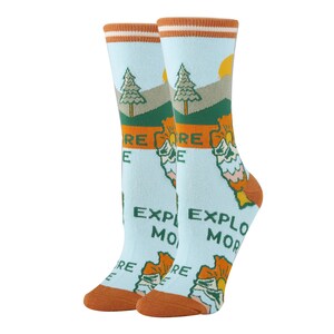 May include: A pair of blue socks with a graphic of a mountain range, trees, and a sun. The socks have orange and white stripes at the top and the text "Explore More" is printed on the socks.