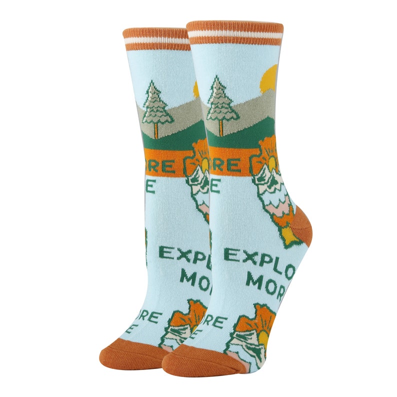 May include: A pair of blue socks with a graphic of a mountain range, trees, and a sun. The socks have orange and white stripes at the top and the text "Explore More" is printed on the socks.