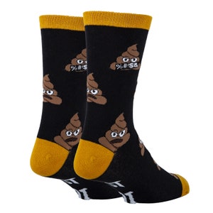 Puede incluir: Un par de calcetines negros con un pu&ntilde;o amarillo. Los calcetines presentan un patr&oacute;n repetido de emojis de caca marrones con caras y el texto "%&#$&" en algunos de los emojis.