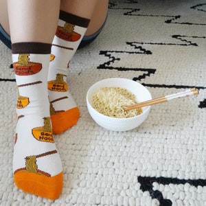May include: White socks with brown cuffs and orange toes feature a ramen bowl graphic with the text "Send Noods." A white bowl of noodles with chopsticks rests on a white and black patterned rug.
