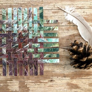 May include: A woven square made of strips of paper with a green and brown marbled pattern. The square is on a wooden surface with a pine cone and a feather.