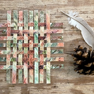 May include: A woven paper coaster with a floral pattern in shades of green, brown, and pink. The coaster is made of strips of paper that are woven together to create a grid pattern.