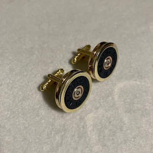 Puede incluir: Un par de gemelos dorados con un centro negro y la palabra "REMINGTON" impresa en el borde. Los gemelos tienen un diseño circular con un borde dorado y un cierre de poste.