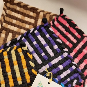 May include: Four woven pot holders in different colors and patterns. The pot holders are brown and beige, purple and black, yellow and black, and blue and black. A tag is attached to the pot holders that says "Thank You Tuesday Afternoons" with a website address.