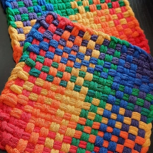 May include: Two woven pot holders in rainbow colours. The pot holders are made from a thick, textured yarn and are square shaped.