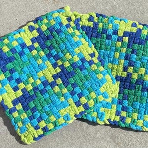 May include: Two square potholders featuring a woven grid pattern made from thick yarn in shades of lime green, bright turquoise, and royal blue. The textured fabric displays a consistent interlocking weave design throughout both square items.