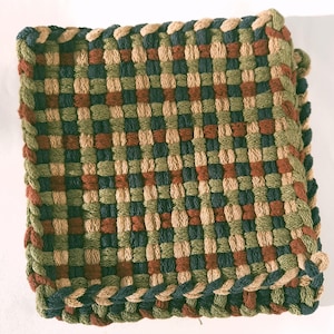 May include: Square woven trivet in a checkerboard pattern. The trivet features a mix of colors including green, brown, and navy blue. The edges are finished with a braided trim in coordinating colors. This item is suitable for kitchen use.