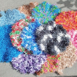 May include: A collection of round, textured scrubbers made from shaggy, multi-coloured synthetic yarn. The scrubbers feature various colour combinations including solid blue, black with white accents, bright green, orange, pink, and beige tones.